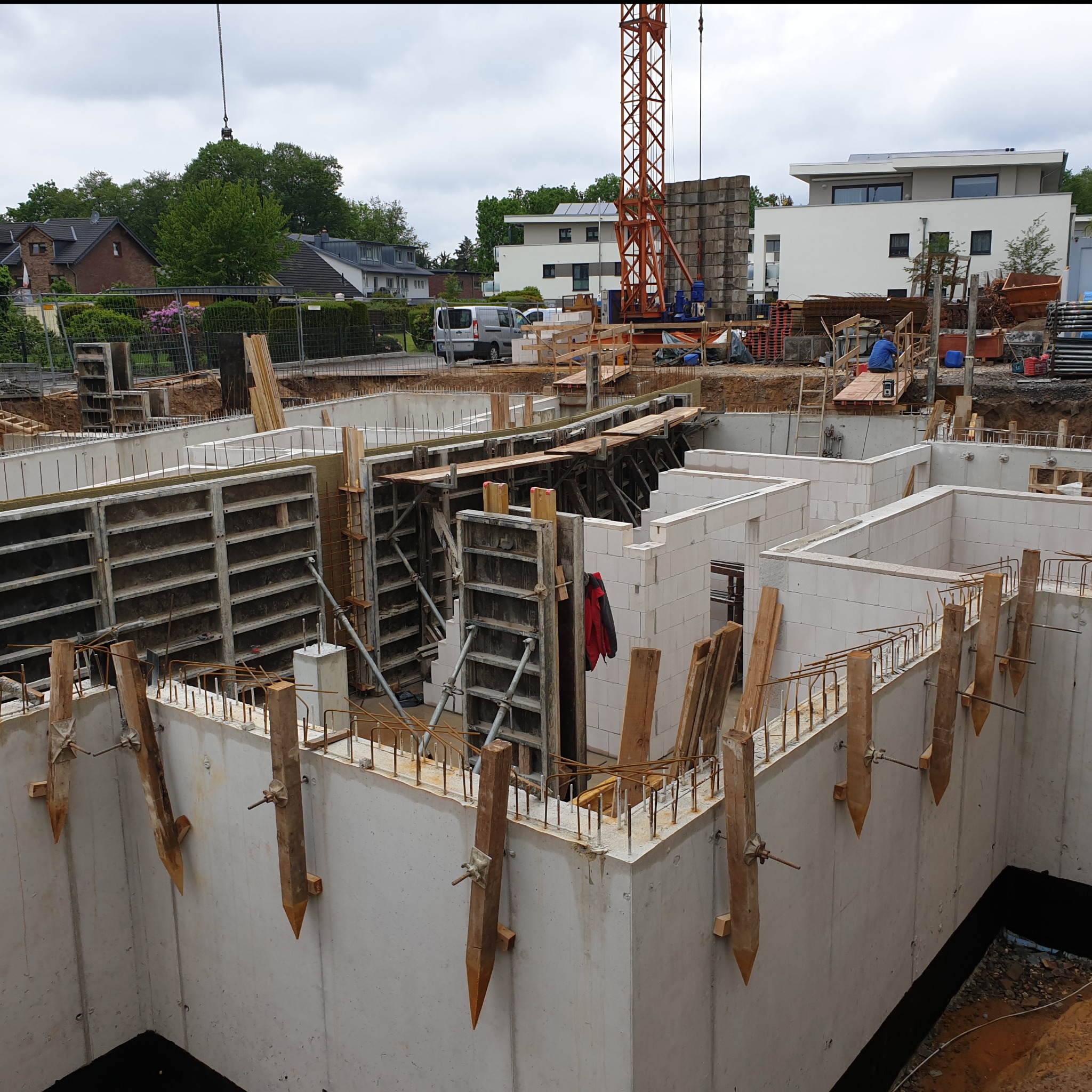 Budowa fundamentów domu: betonowe ściany z drewnianymi podporami, widoczne zbrojenia i elementy szalunkowe, w tle żuraw budowlany i zabudowa mieszkalna.