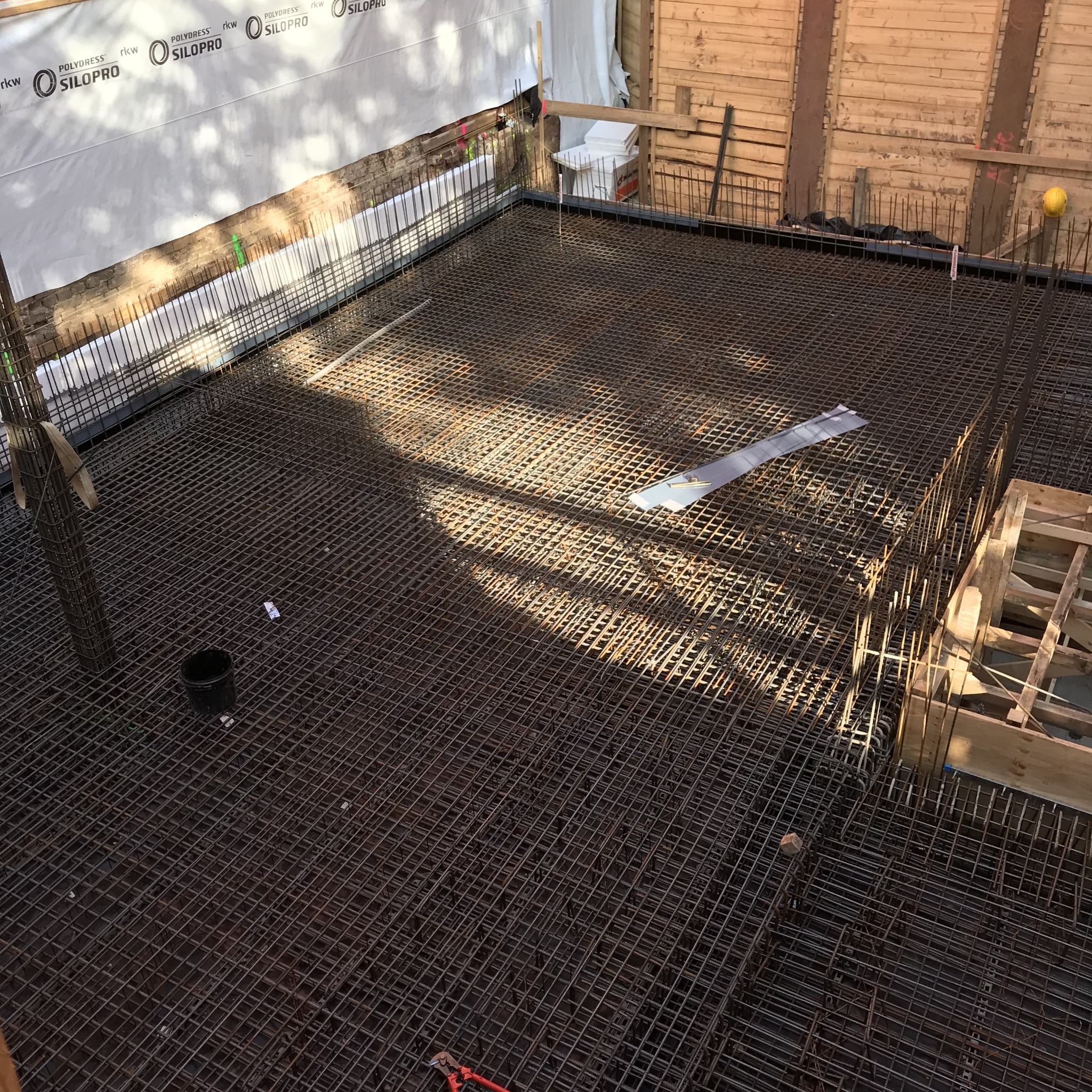 Siatka zbrojeniowa na fundamencie pod budowę domu, widoczne narzędzia i elementy konstrukcyjne, zabezpieczona folia w tle.