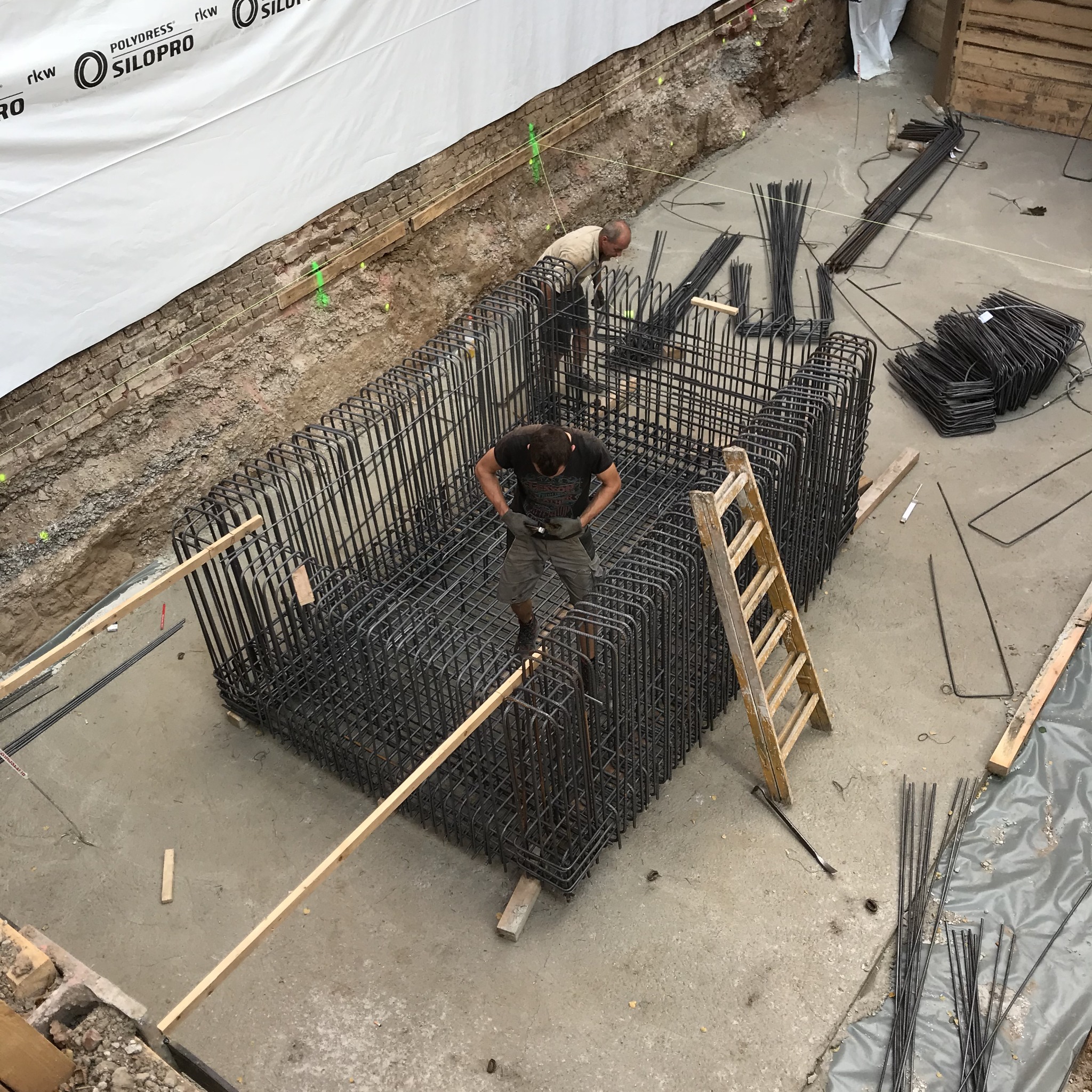 Zbrojenie fundamentów pod budynek, widok z góry na plac budowy z dwoma pracownikami, drewnianą drabiną i stertą prętów zbrojeniowych.