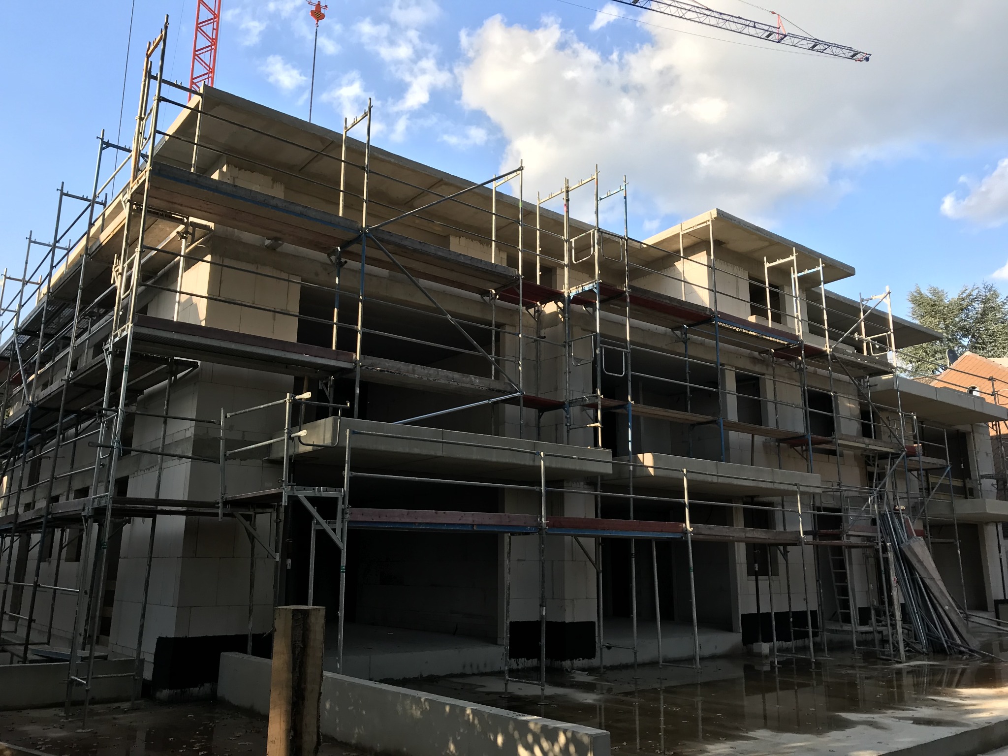 Wielopiętrowy budynek w trakcie wznoszenia z białych bloczków, otoczony rusztowaniami, z widocznym żurawiem budowlanym na tle błękitnego nieba z chmurami.