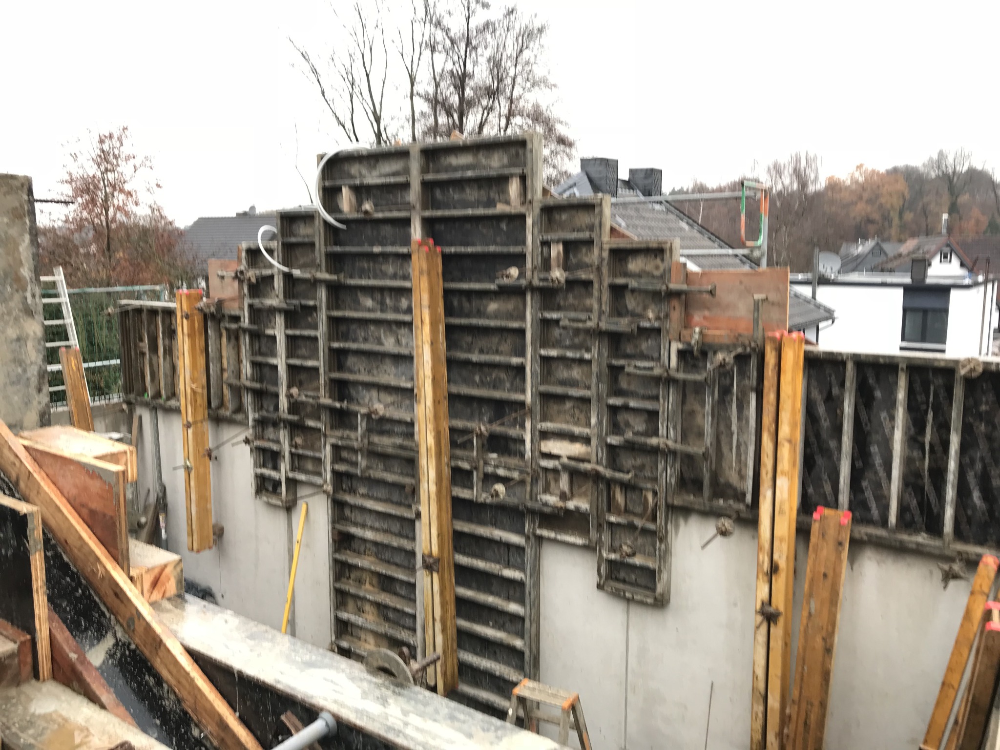 Szare szalunki na betonowych ścianach fundamentów domu w trakcie budowy, drewniane stemple podtrzymujące konstrukcję, drabina oparta o ścianę, widok na okoliczne domy i drzewa.