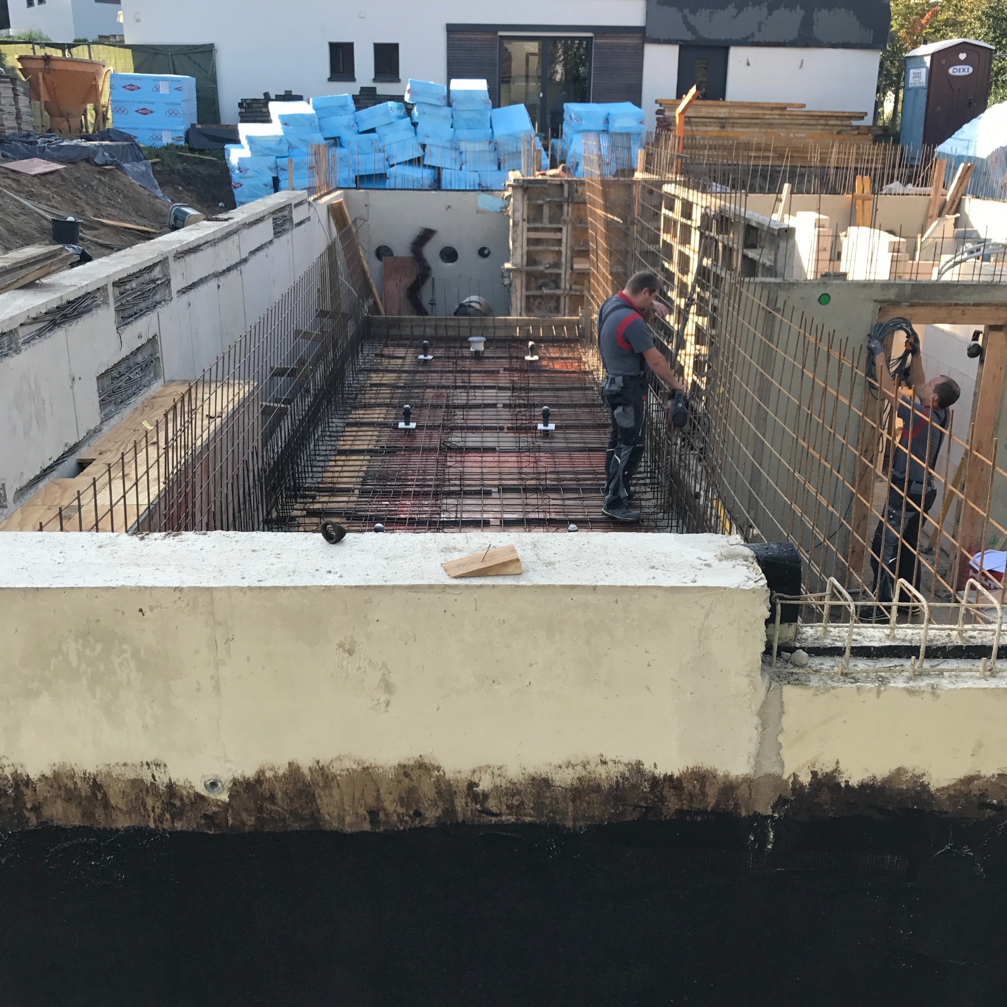 Budowa fundamentów: betonowa konstrukcja zbrojona stalą, pracownicy montujący elementy zbrojenia, widoczne izolacja przeciwwilgociowa i materiały budowlane w tle.