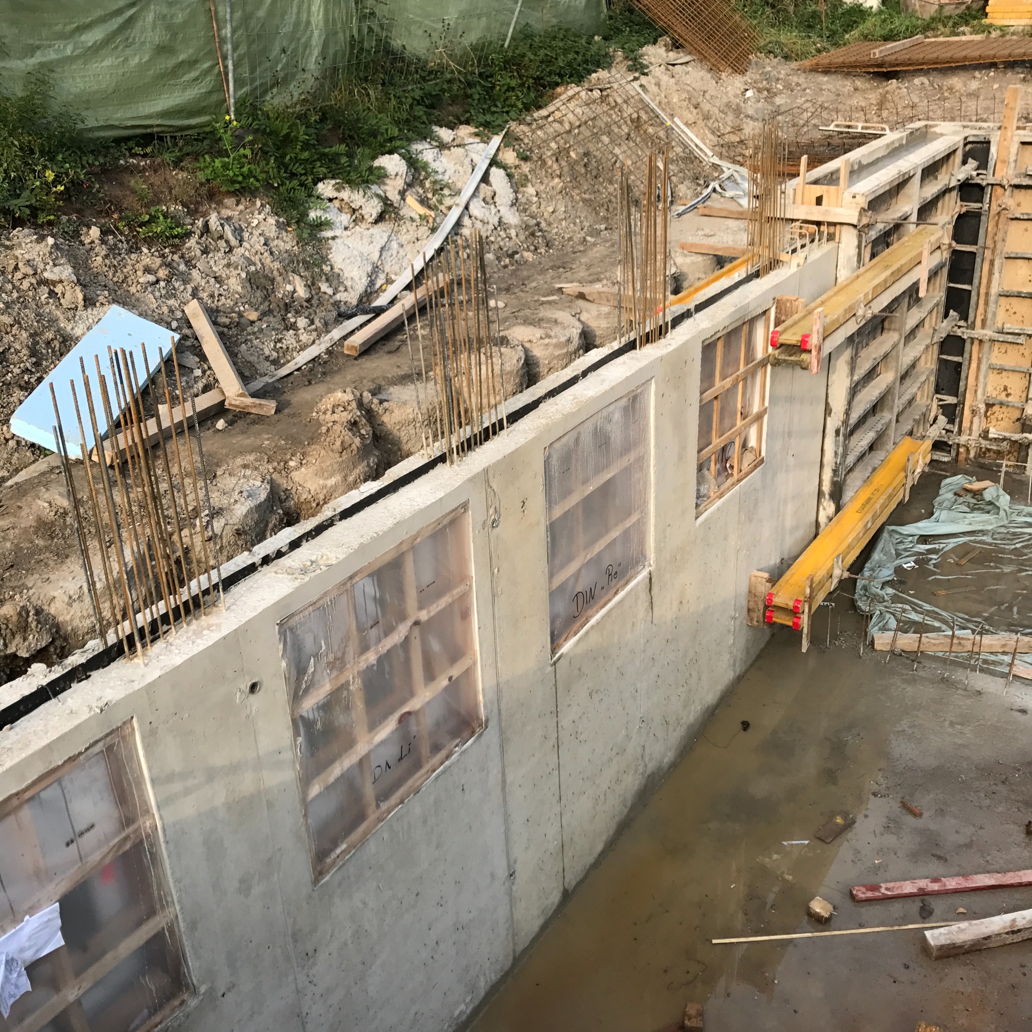 Betonowa ściana fundamentowa z otworami okiennymi zabezpieczonymi folią, stalowe pręty zbrojeniowe wystające z góry, szalunki drewniane po prawej stronie, teren budowy w trakcie prac ziemnych.