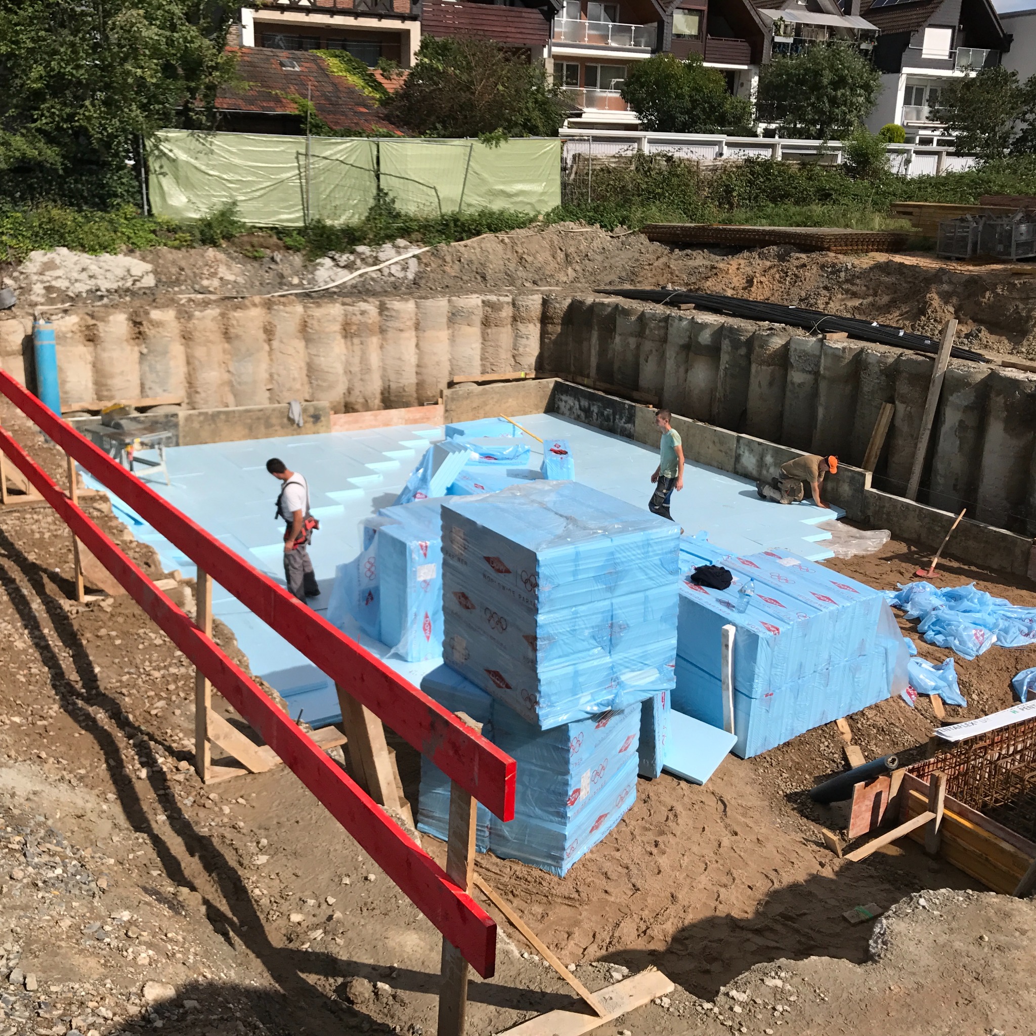 Fundamenty domu jednorodzinnego w trakcie ocieplania styropianem, widoczni dwaj pracownicy budowlani oraz stosy niebieskiego styropianu w folii, zabezpieczone wykopaliska w tle.