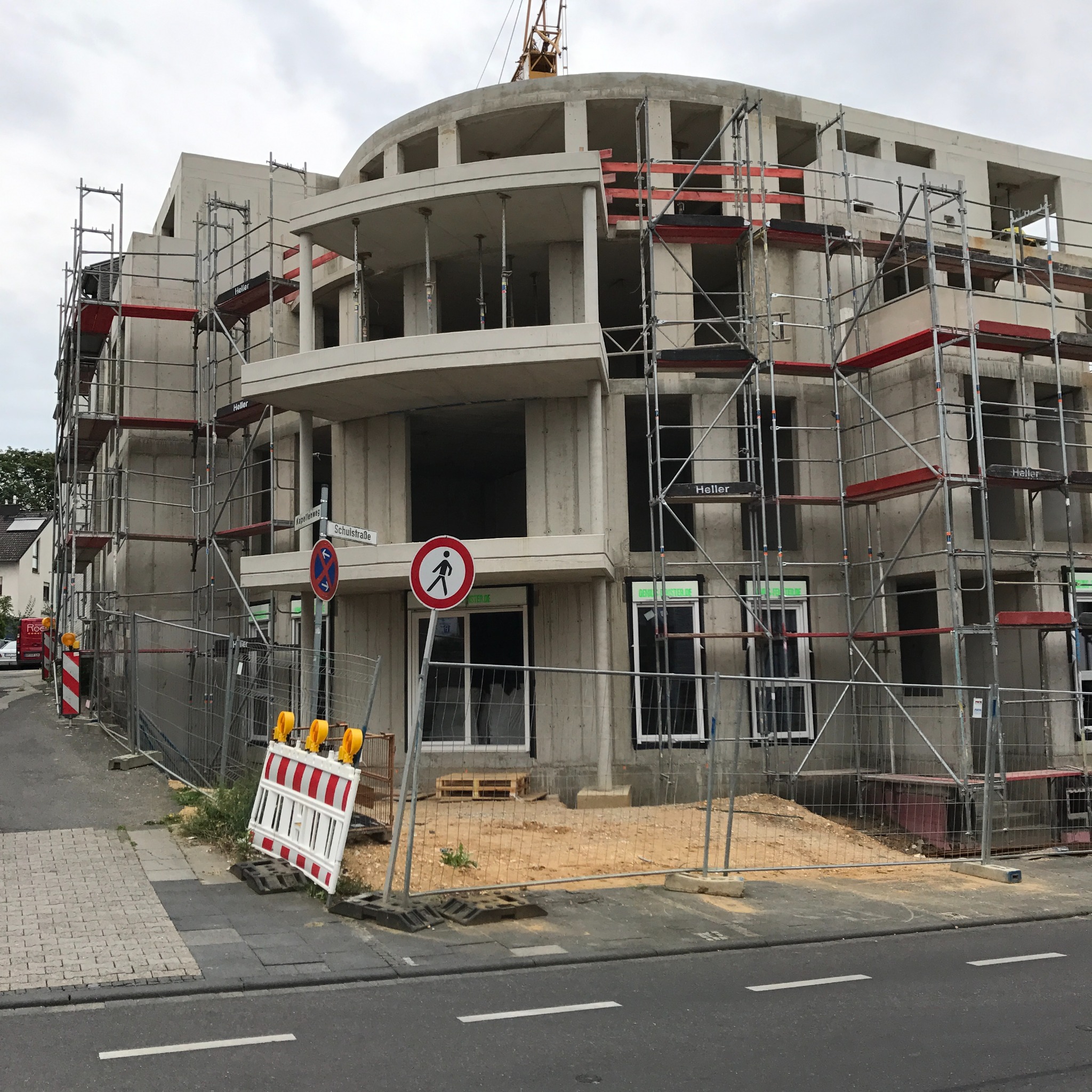 Wznoszony wielopiętrowy budynek z betonu komórkowego, otoczony rusztowaniami i ogrodzeniem budowlanym z żółtymi lampami ostrzegawczymi, widoczne znaki drogowe.