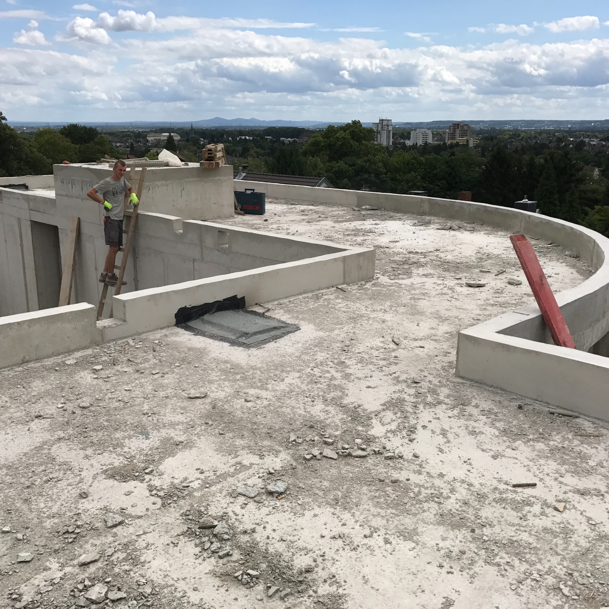 Budowa domu murowanego: robotnik na drabinie na dachu w trakcie prac, widok na panoramę miasta w tle.