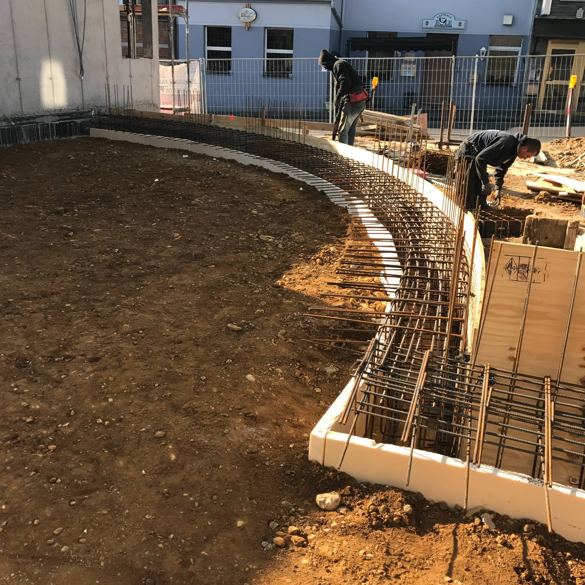 Zbrojenie fundamentów o łukowatym kształcie, dwóch pracowników w czarnych bluzach przy pracy, widok na plac budowy z ziemią i elementami konstrukcyjnymi, budynek w tle.