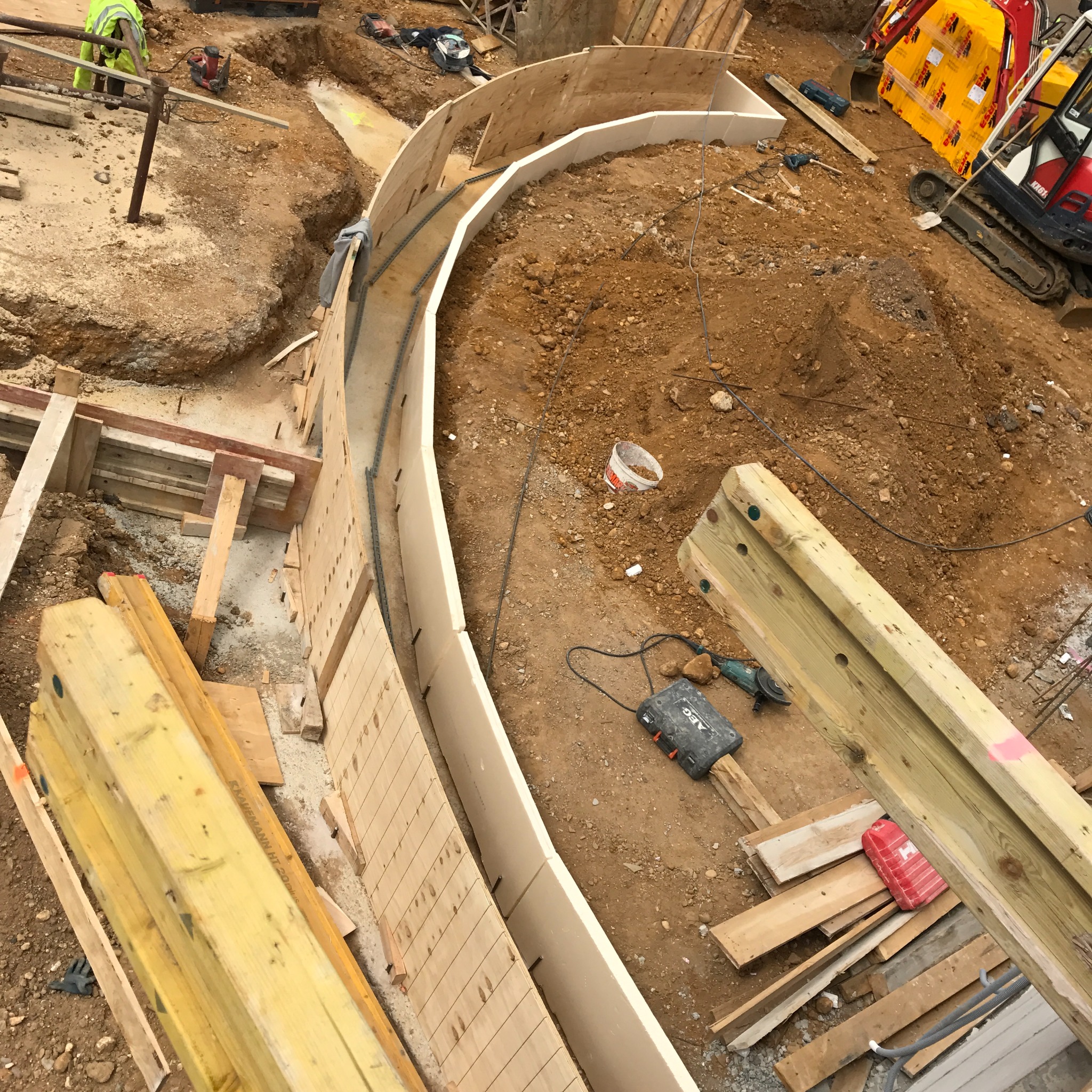 Przygotowanie fundamentów pod budynek z zakrzywionymi ścianami, widoczne szalunki i zbrojenie w wykopie, sprzęt budowlany w tle.