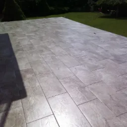 Nowa, błyszcząca posadzka z szarych, prostokątnych płytek imitujących kamień, ułożonych na zewnątrz, z widocznym cieniem budynku i zielonym trawnikiem w tle.