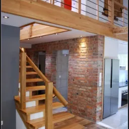 Jasne drewniane schody z białymi podstopnicami w nowoczesnym wnętrzu z ceglaną ścianą i drewnianą antresolą z metalowymi barierkami.