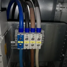 Podłączenie przewodów elektrycznych oznaczonych L1, L2, L3 i N do listwy zaciskowej wewnątrz metalowej obudowy, widoczne oznaczenia faz na żółtych etykietach.