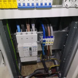 Wnętrze rozdzielnicy elektrycznej z zainstalowanymi bezpiecznikami, przewodami oznaczonymi kolorami i złączkami, widok z dołu w trakcie montażu.