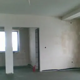 Wnętrze w trakcie prac wykończeniowych: ściany pokryte świeżym tynkiem gipsowym, widoczne otwory okienne i drzwiowe, na podłodze minimalne zabrudzenia po pracach tynkarskich. W tle okrągłe okno.