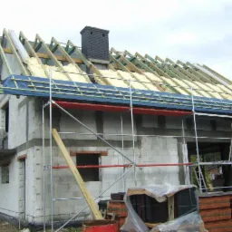 Surowy stan domu z białych bloczków, widoczne rusztowania, więźba dachowa z ociepleniem i komin z cegły klinkierowej, przygotowane dachówki na ziemi.