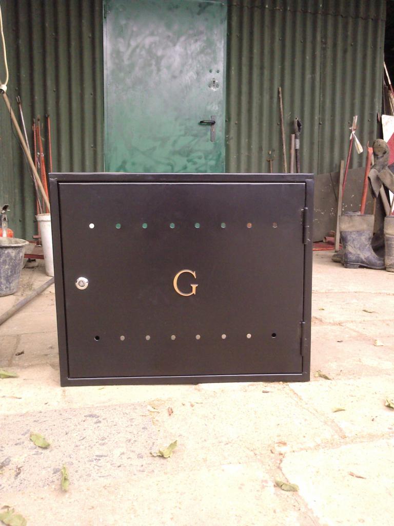 Czarna szafka gazowa z wygrawerowaną literą 'G', umieszczona na zewnątrz budynku z blachy falistej i zielonymi drzwiami.