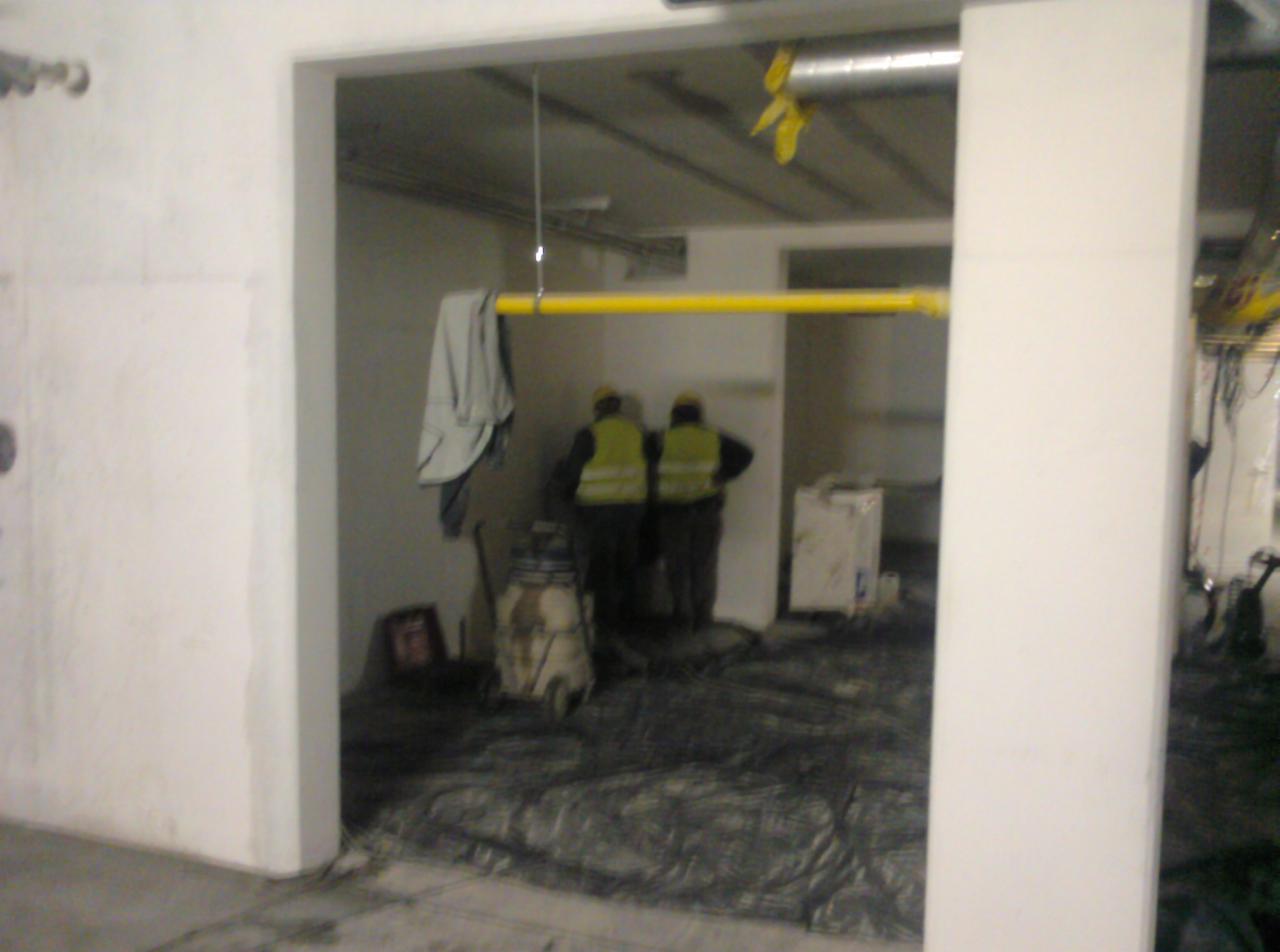 Wnętrze w trakcie prac instalacyjnych: dwaj pracownicy w żółtych kamizelkach stoją obok żółtego rurociągu zawieszonego pod sufitem, widoczne narzędzia i zabezpieczenia podłogi.