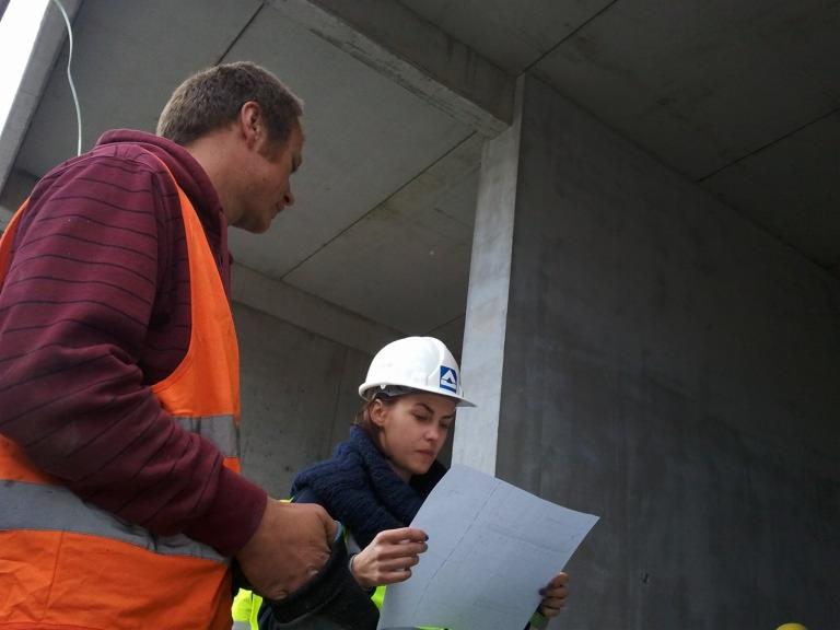 Dwóch inżynierów w kamizelkach odblaskowych i kaskach analizuje plan instalacji przy betonowej ścianie w surowym stanie.