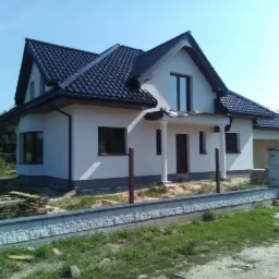 Gotowy, dwukondygnacyjny dom z ciemną dachówką, białymi ścianami i elementami wykończeniowymi w kolorze brązowym, otoczony niskim, kamiennym murem i częściowo ogrodzeniem z drewnianych desek.