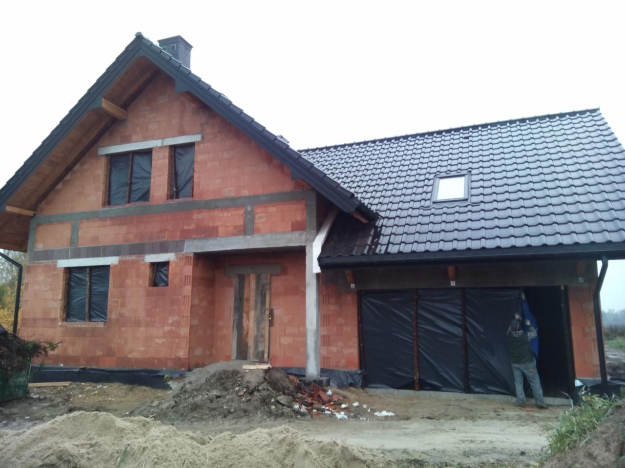 Surowy stan domu z czerwonej cegły, z czarną dachówką i prowizorycznie zasłoniętymi oknami, widoczny pracownik przy garażu, ziemia wokół budynku.