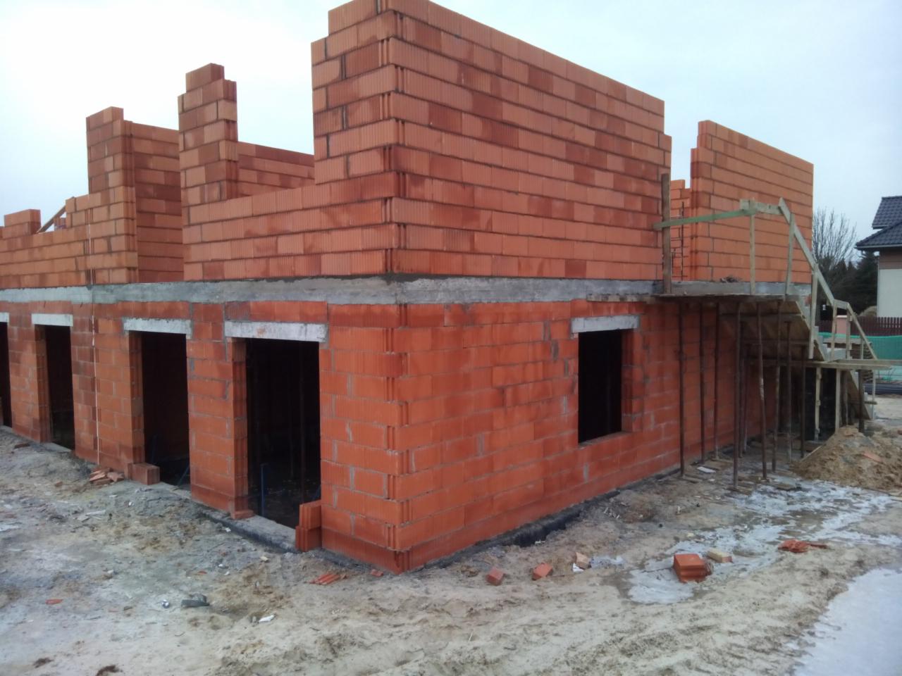 Surowy stan domu w trakcie budowy z czerwonej cegły, widoczne otwory okienne i drzwiowe, częściowo wzniesione ściany, szare niebo w tle.