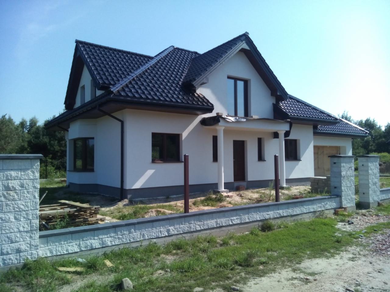 Gotowy, dwukondygnacyjny dom z ciemną dachówką, białymi ścianami i elementami wykończeniowymi w kolorze brązowym, otoczony niskim, kamiennym murem i częściowo ogrodzeniem z drewnianych desek.