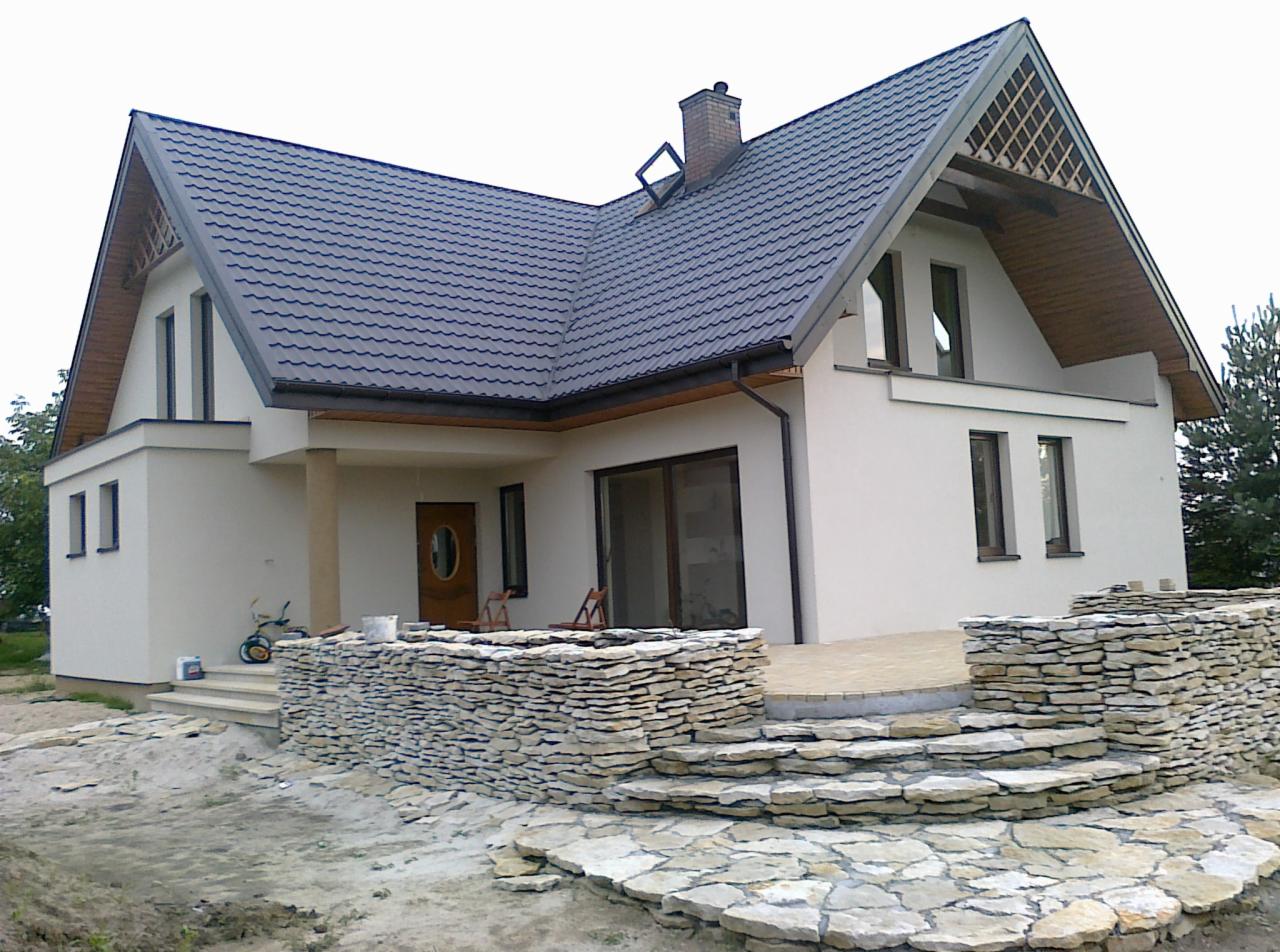Nowoczesny dom z szarą dachówką i kamiennym tarasem w trakcie budowy, widoczne materiały budowlane i narzędzia.