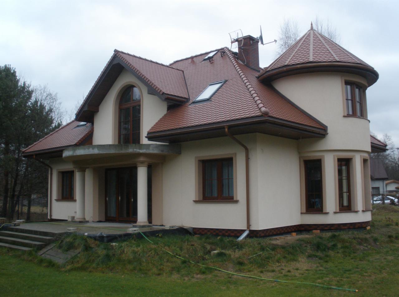 Nowo wybudowany, jasny dom z brązową dachówką i okrągłą wieżyczką; widoczne kolumny przy wejściu i schody prowadzące do budynku.