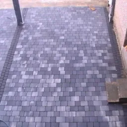 Ułożona kostka brukowa w odcieniach szarości, z ciemniejszym obramowaniem i widocznymi narzędziami brukarskimi przy krawędzi.