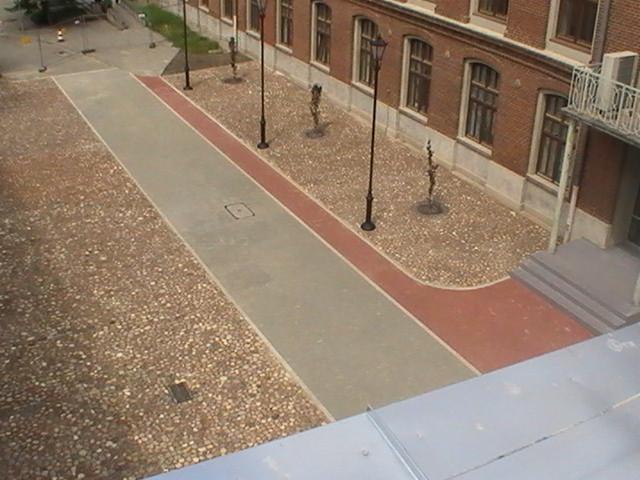 Dziedziniec Politechniki Łódzkiej po renowacji, widok z góry: szara, betonowa ścieżka z czerwoną obwódką, otoczona drobnymi kamieniami, oświetlona stylizowanymi latarniami. Widoczny fragment...