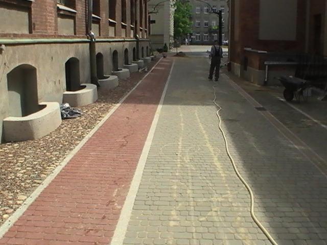 Nowo ułożona dwukolorowa kostka brukowa w Łodzi, widok z perspektywy chodnika wzdłuż budynku Politechniki Łódzkiej, w tle pracownik z wężem.