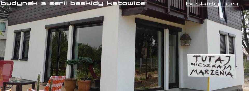 Biały budynek z serii Beskidy Katowice z ciemnymi roletami i napisem 'Tutaj mieszkają marzenia'. Widok na elewację z oknami i fragment ogrodu.