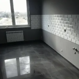 Wykończona łazienka z szarą podłogą z płytek i białymi, błyszczącymi płytkami na ścianie do połowy wysokości. Widoczne przyłącza wodne i grzejnik pod oknem.