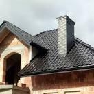 Fragment dachu z ciemnej dachówki ceramicznej i komin z cegły klinkierowej na tle surowej konstrukcji domu w stanie budowy.