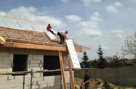 Pracownicy na dachu w trakcie montażu izolacji z białych płyt styropianowych, drewniana konstrukcja dachu, częściowo wymurowane ściany budynku w tle.