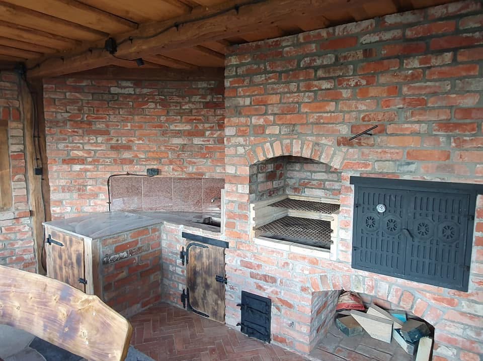 Murowany grill z cegły z drewnianymi elementami, zadaszony drewnianą konstrukcją. Widoczne ruszty, drzwiczki i blat roboczy. Całość utrzymana w rustykalnym stylu.