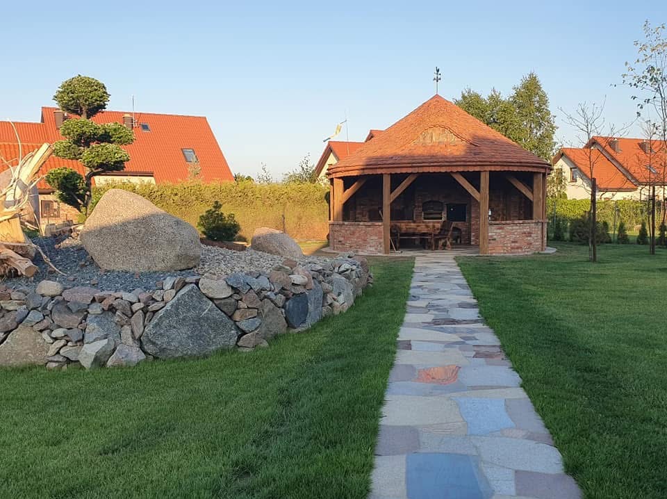 Ogród z altaną z cegły i drewna, kamienny murek z dużymi głazami, ścieżka z płyt kamiennych, trawnik i domy w tle. Słoneczny dzień, spokojna atmosfera.