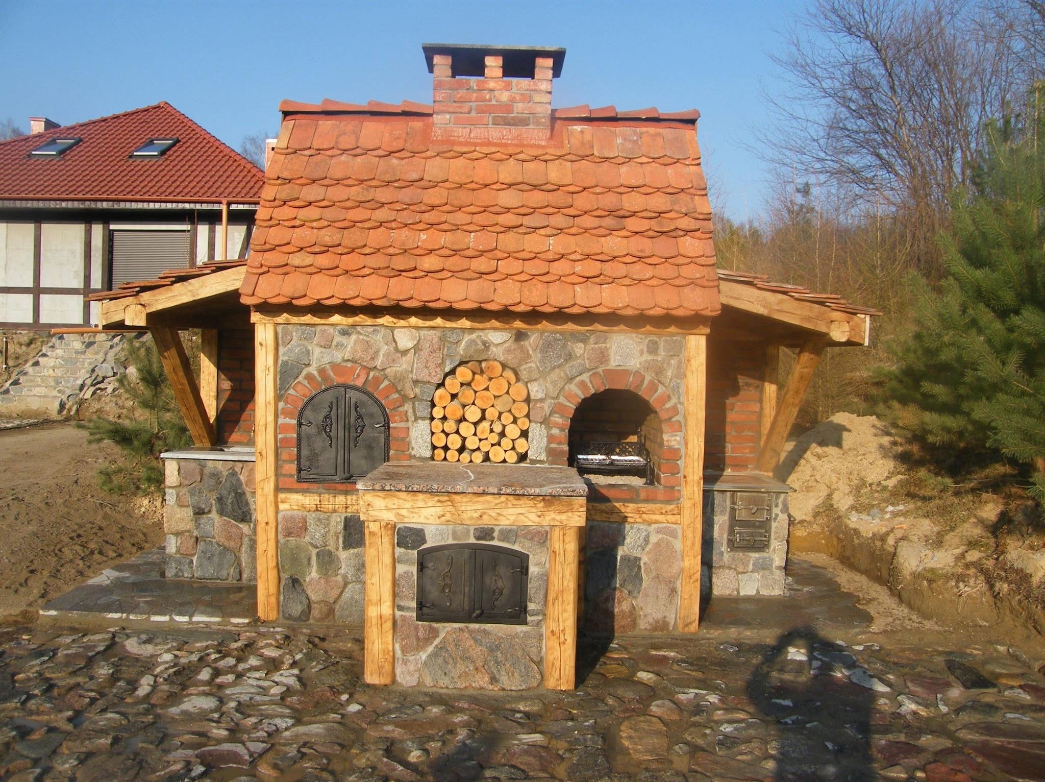 Rustykalny, murowany grill ogrodowy z piecem chlebowym, obłożony kamieniem i cegłą, z drewnianą konstrukcją dachu krytego dachówką. Widoczne drewno na opał.