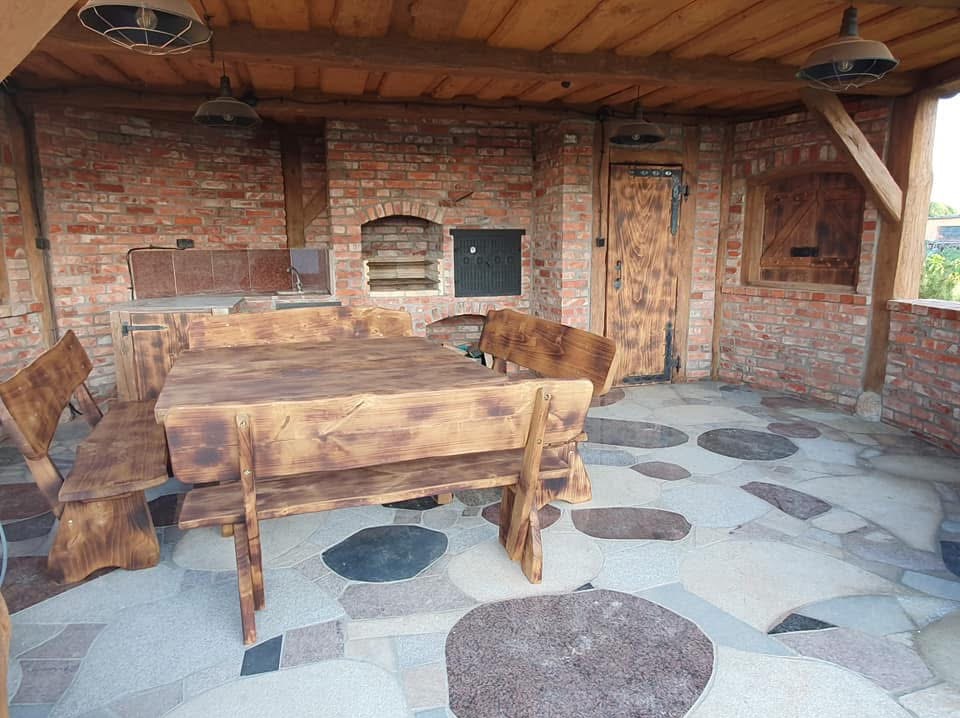 Zadaszony taras z ceglaną zabudową grilla, drewnianym stołem i ławami. Podłoga wyłożona kamieniami o nieregularnych kształtach. Rustykalny styl.