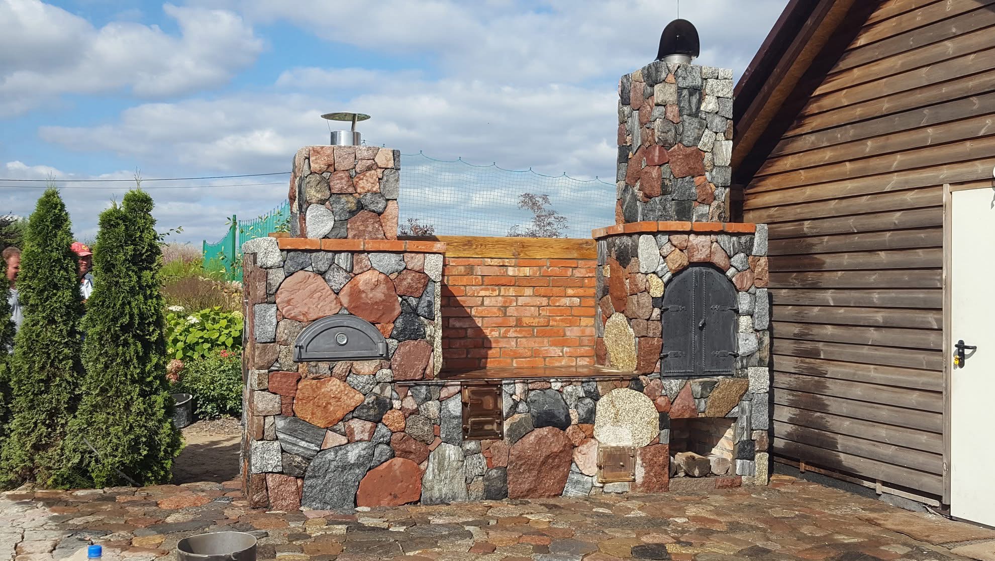 Ogród z kamiennym grillem i piecem chlebowym, z elementami z czerwonej cegły, pod błękitnym niebem. Widoczny fragment drewnianej konstrukcji budynku.