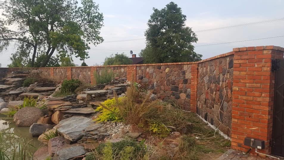 Kamienno-ceglany mur ogrodzeniowy z bramą, otoczony zielenią i skalniakiem z oczkiem wodnym. Widok na fragment ogrodu i dom w tle.