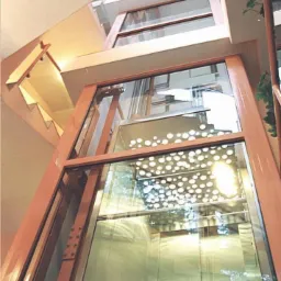 Winda osobowa z przeszkloną kabiną i szybem, widoczna z dołu, z dekoracyjnym perforowanym sufitem w kabinie i elementami konstrukcyjnymi w kolorze terakoty.