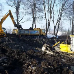 Żółta koparka JCB ładuje ziemię do ciężarówki na placu budowy, obok stoi spychacz marki WOMAG, w tle drzewa i resztki śniegu.