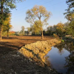 Wykopany staw w naturalnym krajobrazie z drzewami w jesiennych barwach, ziemia świeżo naruszona, woda odbija drzewa.
