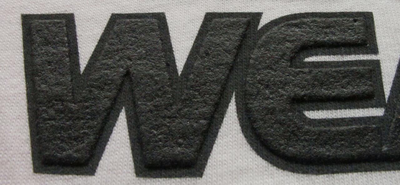 Zbliżenie na fragment białej koszulki z trójwymiarowym, teksturowanym nadrukiem w kolorze grafitowym, przedstawiającym litery 'WE'.