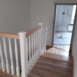 Drewniane schody z białymi balustradami i jasnym drewnianym poręczą, widok z góry na drewniane stopnie i podłogę, w tle otwarte drzwi do remontowanego pomieszczenia z widocznym statywem...