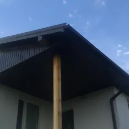 Szary dach z rynnami i podbitką na tle błękitnego nieba. Widoczny fragment ściany budynku z oknami. Drewniany słup podtrzymujący zadaszenie.