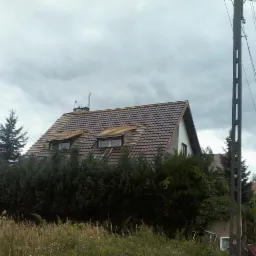 Dom jednorodzinny z brązowym dachem w trakcie wymiany. Widoczne okna dachowe i częściowo zdjęte dachówki. Tło: pochmurne niebo i zieleń.