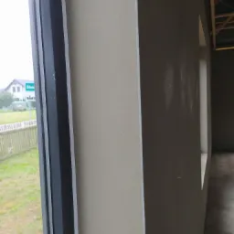 Świeżo nałożony tynk maszynowy na ścianie wewnętrznej, widoczna rama okienna z widokiem na zewnątrz, surowe wykończenie w tle.