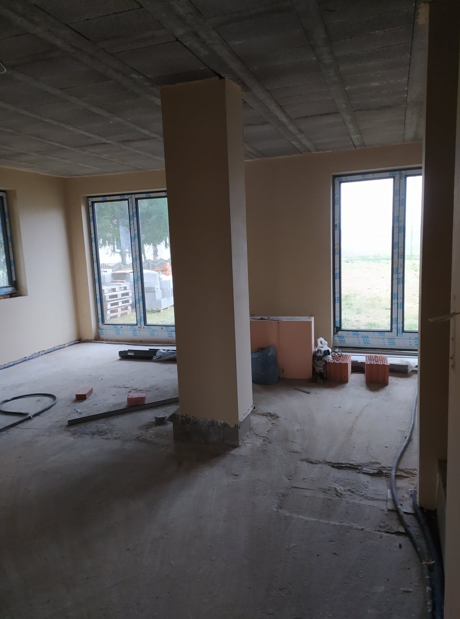 Surowe wnętrze w trakcie prac wykończeniowych: ściany pokryte jasnym tynkiem, betonowa podłoga z widocznymi niedoskonałościami, wystające kable, cegły i materiały budowlane przy oknach. Widoczny...