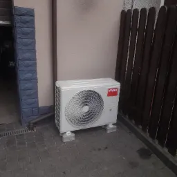Biała jednostka zewnętrzna klimatyzacji marki Vivax, zamontowana na betonowych podstawkach przy ścianie budynku w Kobyłce, widoczna z poziomu brukowanej nawierzchni.